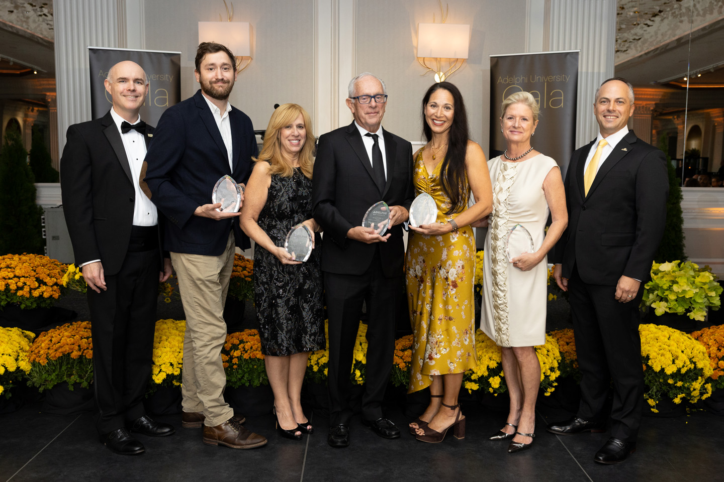 Seven formally dressed individuals stand in a row, five of whom hold plaques of recognition they have received as members of the Bridges to Adelphi Board of Advisors.