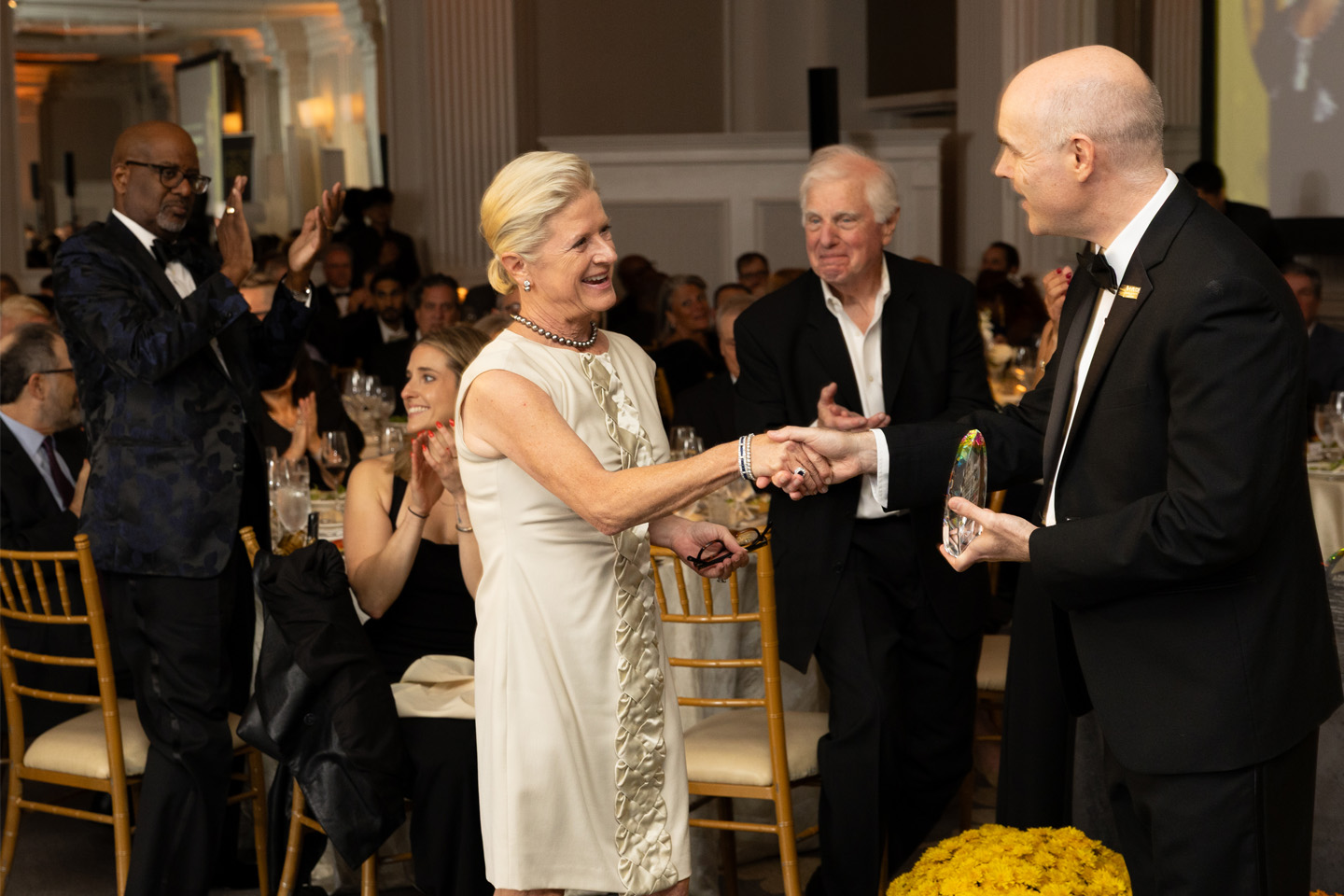 Amidst Gala attendees in formal wear, Adelphi University Board of Trustees Chair Katy Carey stands to receive recognition from Interim President Christopher Storm, PhD.