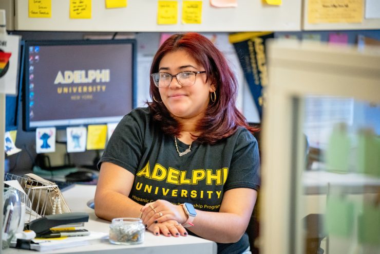 Priscilla sitting at her desk with SafeZone and post-its surrounding her. Wearing and Adelphi University shirt.