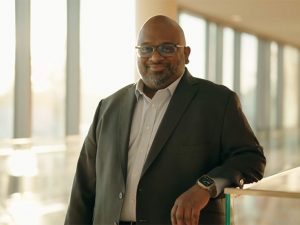 A well-dressed man with a proud smile poses in the hallway of the Nexus building, home of Adelphi's national ranked nursing program.