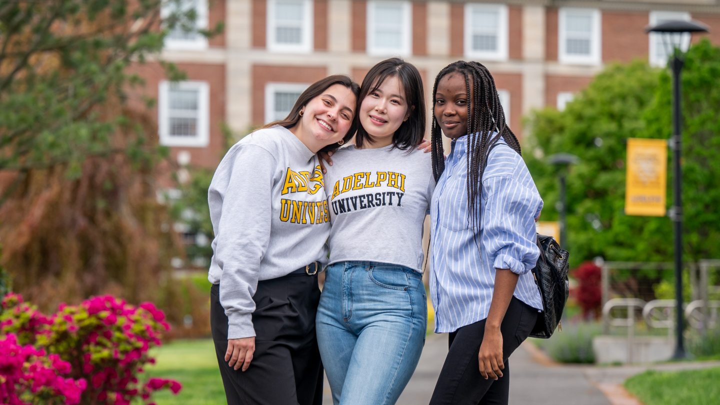 Three students wearing Adelphi Unievsity sweatshirts stand in front of Levermore Hall on campus.