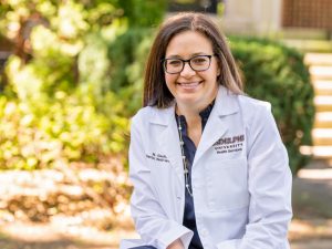 A woman with brown hair and glasses is smiling. She is wearing a white lab coat with the words "Adelphi University Health and Wellness" on the right lapel. Behind her are the grass and trees of the Adelphi campus.