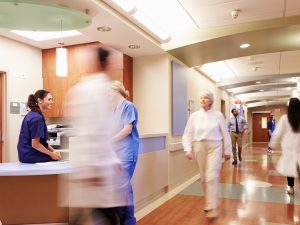 Two female nurses in blue scrubs chat by the nurses station while five hospital staffers walk up and down the brightly lit corridor. 