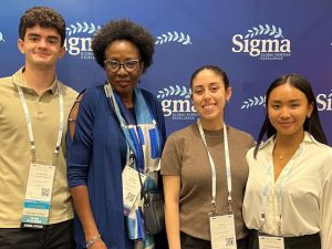 One male and three female students wearing neck lanyards stand in front of a wall labeled Sigma Global Nursing Excellence.