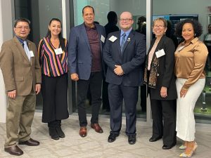A group of six professionally dressed individuals, three men and three women, stand side by side indoors, posing for a photo. They are smiling and name tags are visible on each person. The setting features a modern office environment with glass walls, reflections and a conference room visible in the background.