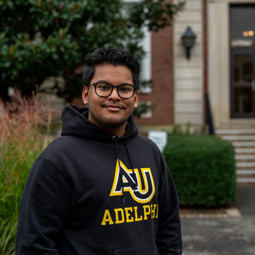A confident male Adelphi University student stands in front of Levermore Hall wearing an Adelphi hoodie.