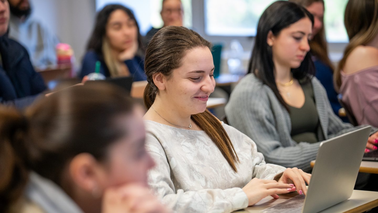 Smiling student typing notes in classroom at Adelphi University.