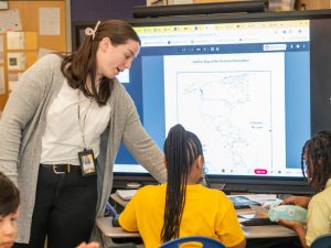 A female teacher leans over a desk to assist two students in a classroom while a map of the Western Hemisphere is displayed on a large digital touchscreen monitor behind them