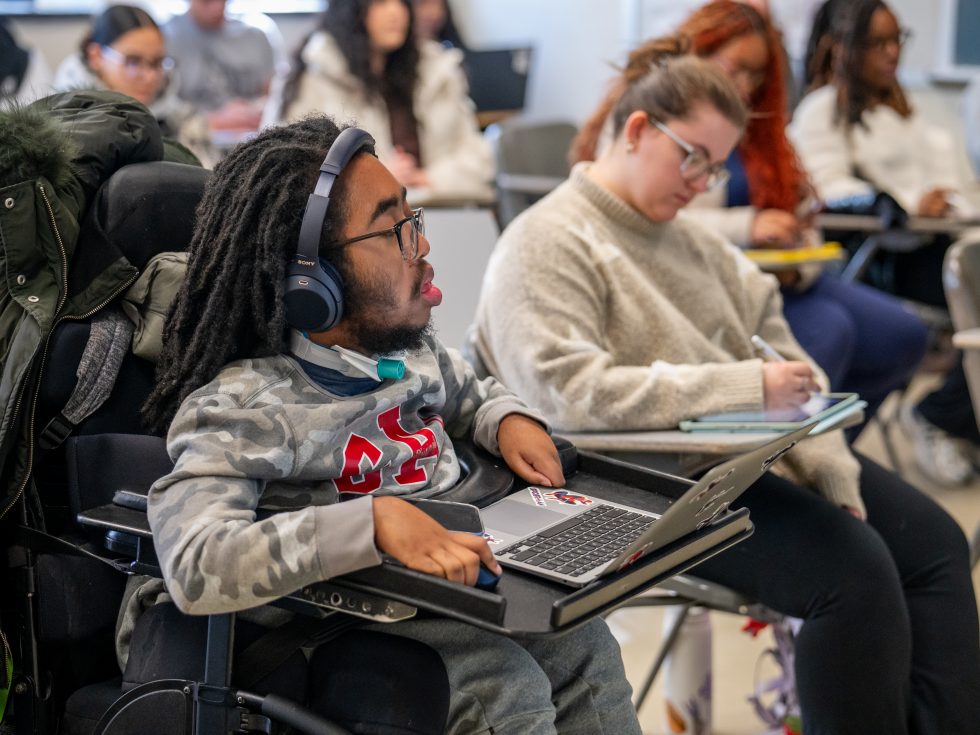 Adelphi University student in class taking notes on laptop while seated in a motorized chair.