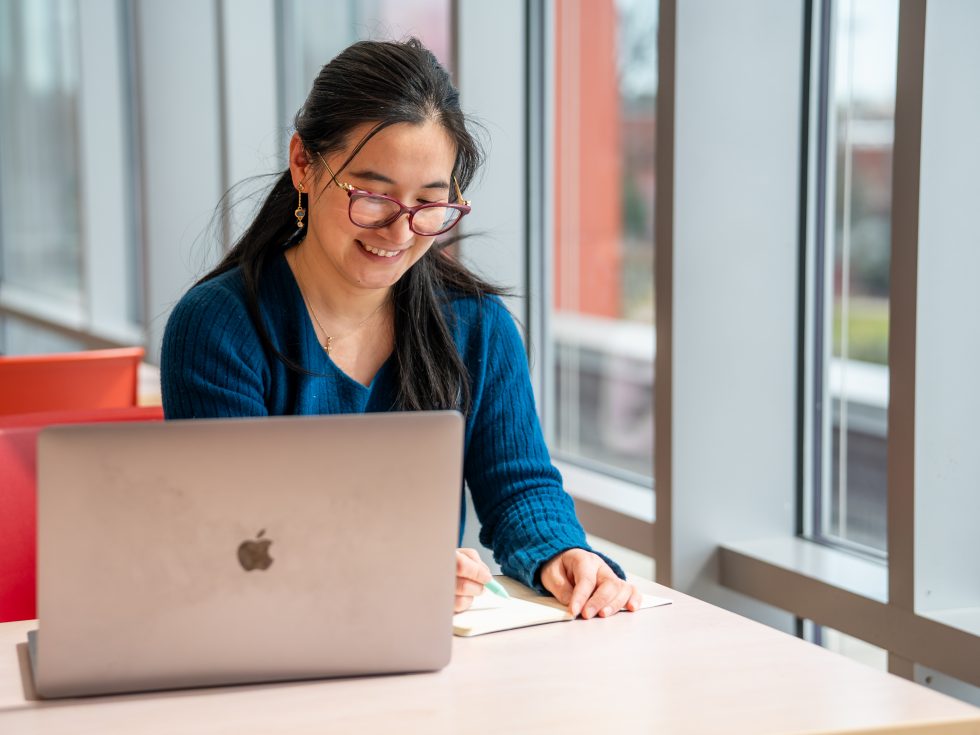 Professor Yiqi Zhu in the Nexus Building writing on her laptop.