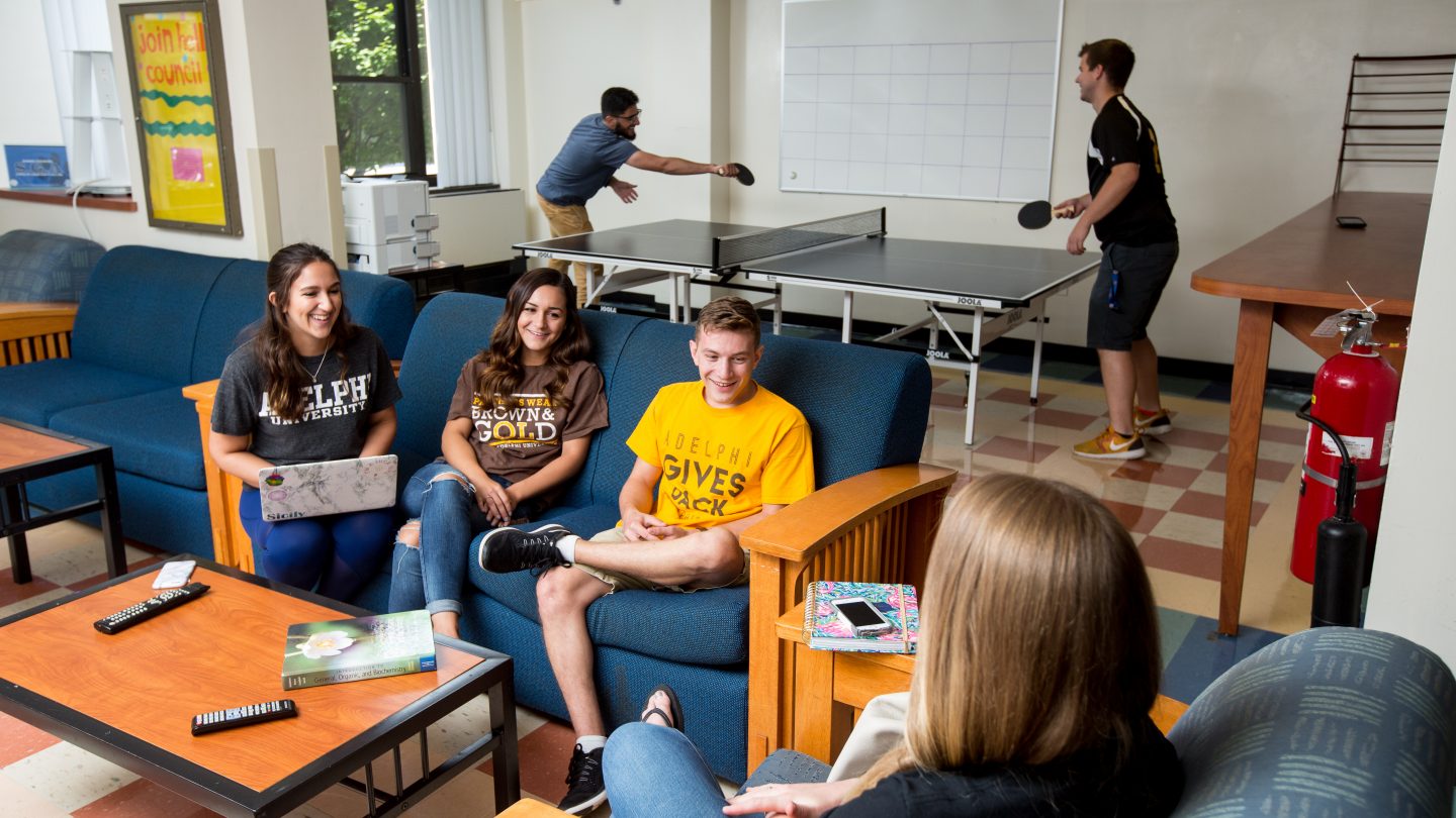 Residential Life on Adelphi University's campus: Friends playing table tennis and chatting on the lounge couch.