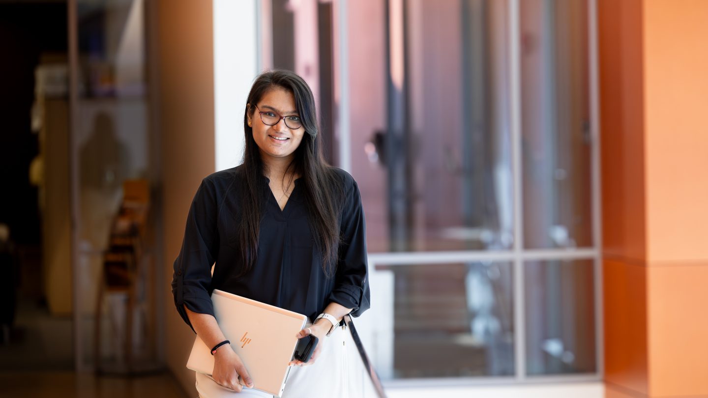 A confident professional Adelphi student holding a laptop in the Nexus Building.