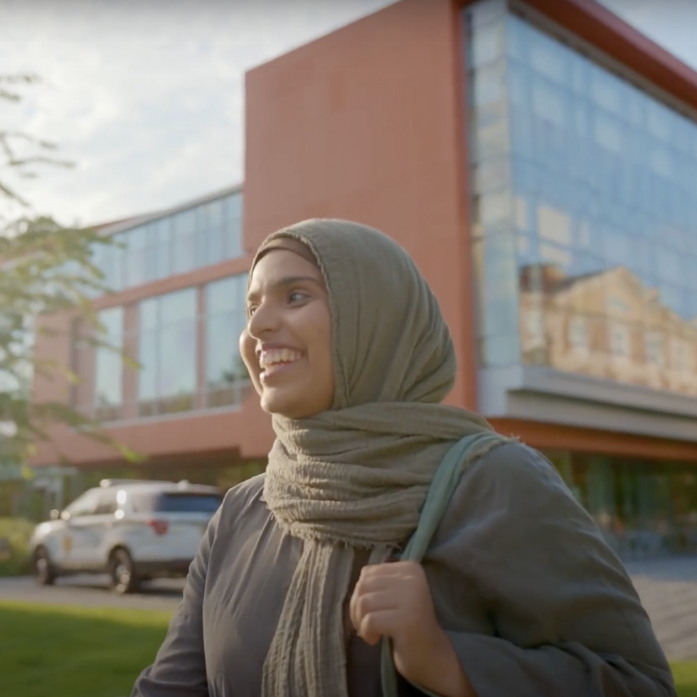 An Adelphi student smiling and walking in front of the Nexus building.