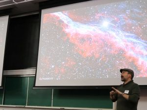 Dr. Coyne stands onstage in front of a projected image of the Veil Nebula.