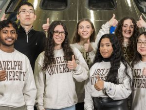 A professor and group of students posing wearing Adelphi University gear giving the thumbs up.
