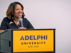 Linda Davis Valdez smiles broadly while giving her address. She is standing before a microphone on a lectern with a gold sign saying Adelphi University, New York.