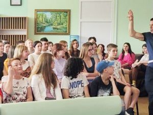 Professor Mirra, holding a book, stands in front of approximately 30 attentive students in a small but bright classroom.
