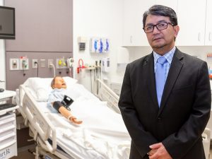 Professor Pajarillo stands in the College of Nursing and Public Health's simulation lab, a hospital-like setting for nurse training. Behind him is a manikin in a hospital bed.