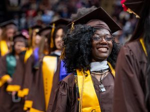 A line of students in graduation gear files into Commencement; one student smiles proudly.