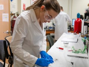 A female research in a lab coat and safety glasses performs research in an Adelphi science lab.