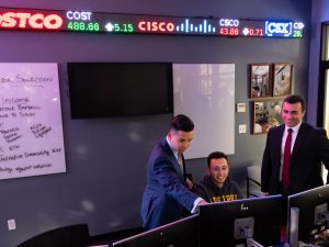 Three students gather around a computer terminal in the trading room. A digital stock ticker and a whiteboard are on the wall behind them.