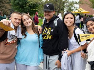 Five students, one wearing an Adelphi sweatshirt, and another holding an Adelphi pennant, pose for the camera on the University's quad.