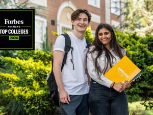 A male and female student in front of a campus building.  The woman holds an Adelphi folder.