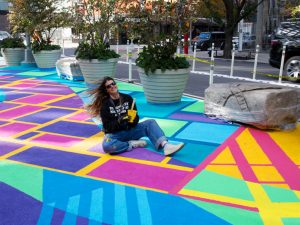Mural artist Cara Lynch ’12 sits on a colorful painted street, paintbrush and paint can in hand, her hair blowing in the wind, smiling broadly. 