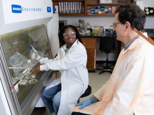 A female student wearing lab gear works with petri dishes while a professor observes