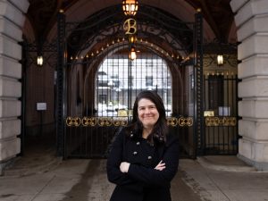 Dr. Provost, in a black winter coat with her arms crossed, stands before the gate to the apartment building's grand entrance.
