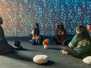 Hoffner sits on a pillow on the floor facing four seated students. In the background, the center's wall twinkles with dozens of tiny lights. The lighting in the center is muted and peaceful.