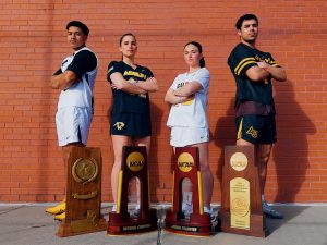 The student-athletes, wearing their teams' uniforms, stand with their arms folded before a brick wall and behind a quartet of national championship trophies.