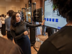A female student presents her work on a video screen as other students stand by.