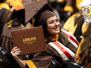 Halpert, in cap and gown, displays her diploma while seated in a row with fellow graduates.