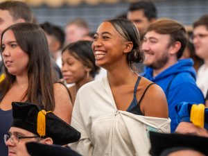 Smiling students amid the crowd of fellow first-year classmates, with faculty members in cap and gown seated in front of them