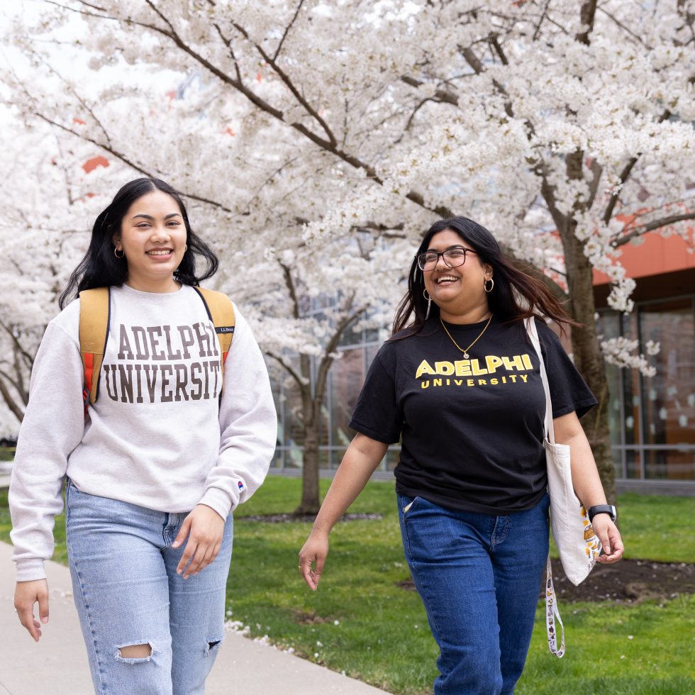 Two Adelphi students walking on campus in the spring with trees in bloom.