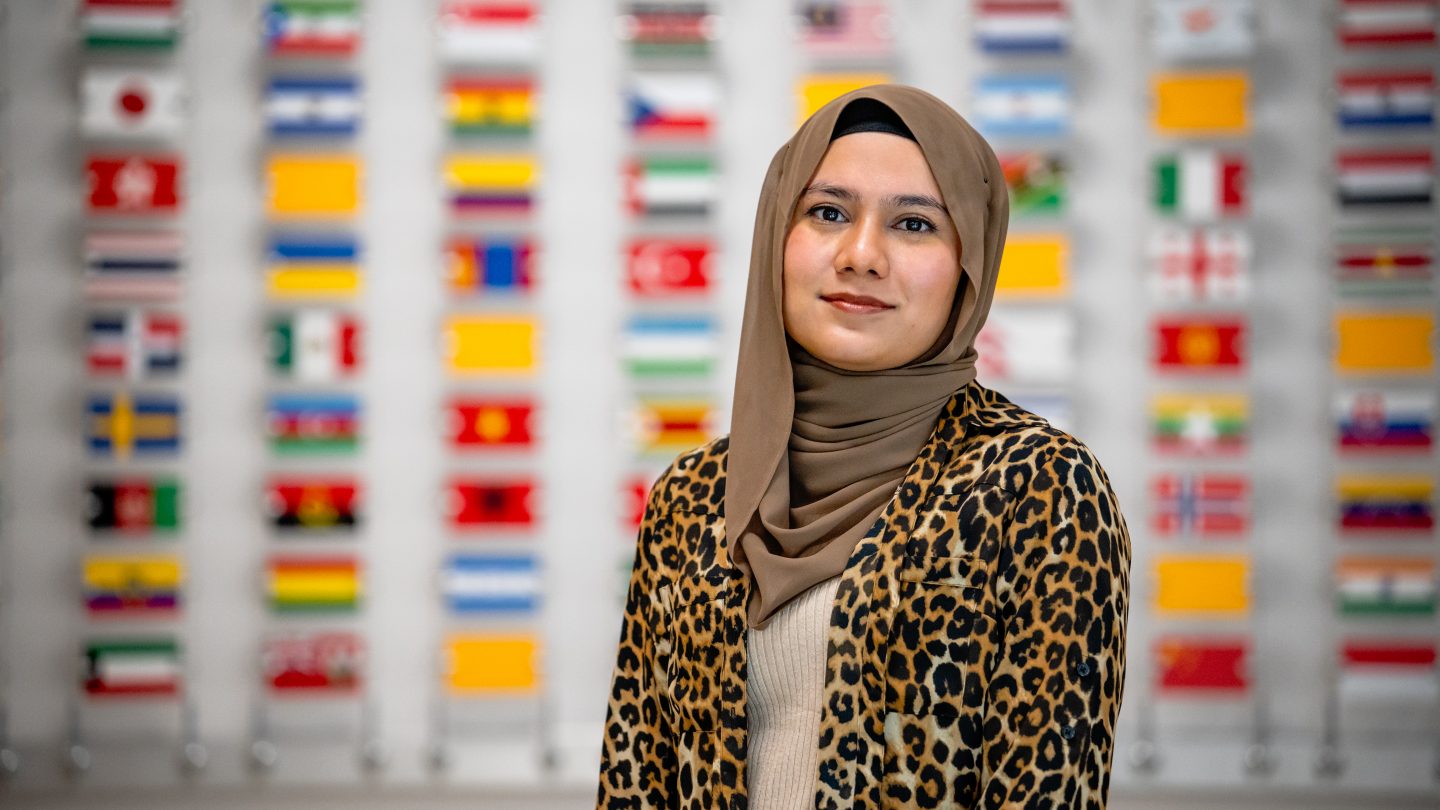Muneera Chowdhury, wearing a hijab, the headscarf worn by Muslim women, stands in front of a wall with the flags of all international students at Adelphi.