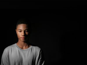 A photo of a young Black man with a somber expression, standing on a stark darkened background.