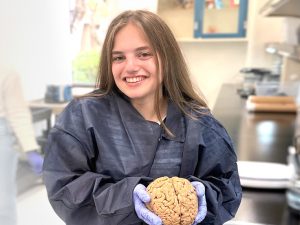 A young woman with long brown hair smiles while wearing a dark blue lab gown and purple gloves. She is holding a human brain specimen in a laboratory setting with lab equipment visible in the background.