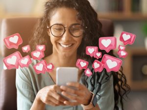 A smiling woman with glasses and long, dark curly hair looks at her smartphone while 3D pink heart notification bubbles float around the screen against a warm background.
