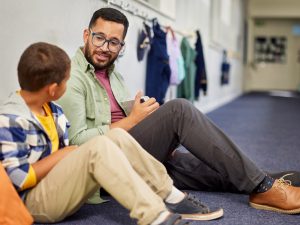 A male school psychologist wearing glasses sits on the carpeted floor of a school hallway, speaking attentively with a young boy sitting beside his orange backpack.