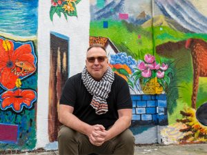 Rob Linné sits on the curb in front of a colorful mural depicting life in El Salvador red hibiscus flowers with a frog one of them, a woman pupusa (the national dish of El Salvador) over an open fire, a llama, a mountain, and a garden wall with a vase of pink tulips on its top.
