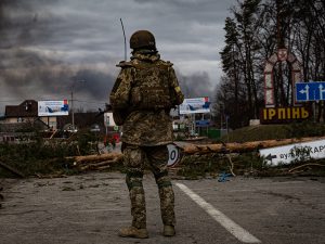  A soldier in camouflage battle fatigues and a helmet stands on a damaged road. He is seen from behind. A tree has fallen across the road, spreading limbs and pine needles across the asphalt. A road sign, in Cyrillic, points the way to Irpin. Billboards can be seen in the distance. Smoke is drifting across a gray sky following shelling by Russian troops.