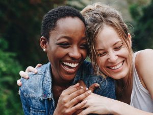 Photo of two women laughing with broad smiles on their face. The women are hugging, with one woman's arm around the other's shoulders. Their heads are touching.