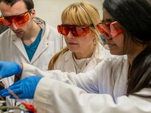 Professor Widera-Kalinowska and two students working in a campus lab. All are wearing white lab coats and orange protective glasses. One of the students is wearing blue exam gloves.