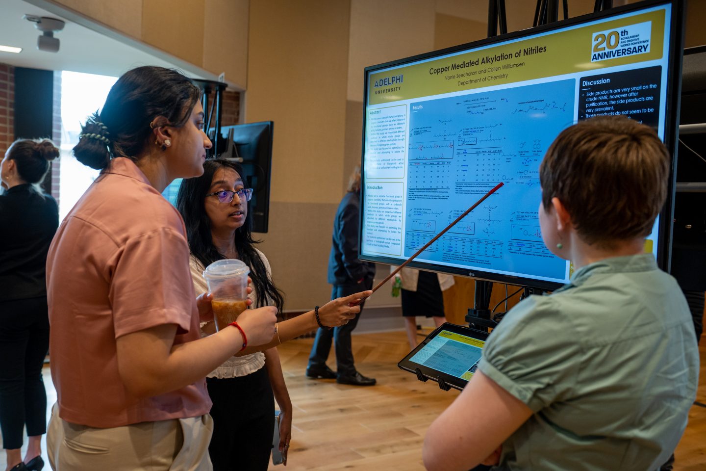 A Department of Chemistry Asian female student aims a pointer at statistics on her team’s Scholarship and Creative Works Conference e-poster, as she explains her topic to two other young women.