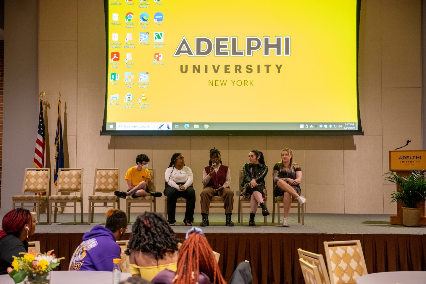 A large video screen is shown above a panel of five young and diverse students who are seated on a stage as they discuss various topics during Research Day.