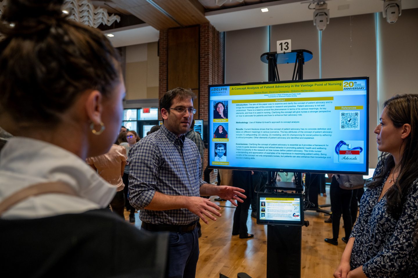 Standing by an e-poster, a white male nursing student wearing eyeglasses, a checkered, long-sleeved shirt and jeans, explains his team’s research to two young women.