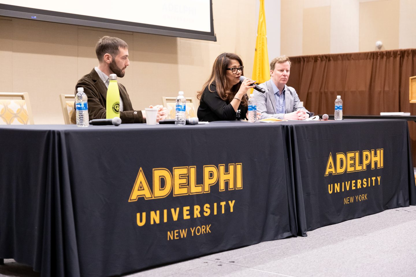 Three adults—two men and one female—are seated at a table draped with Adelphi University signage. She, with long brown hair, eyeglasses and wearing a black blouse, is speaking into a microphone, as the men listen—one with short brown hair and a short beard, wearing a brown jacket and striped shirt, and the other with light brown hair, a pale gray suit jacket and light blue shirt. Four bottles of water are on the table, with a large video screen hanging in the background.  