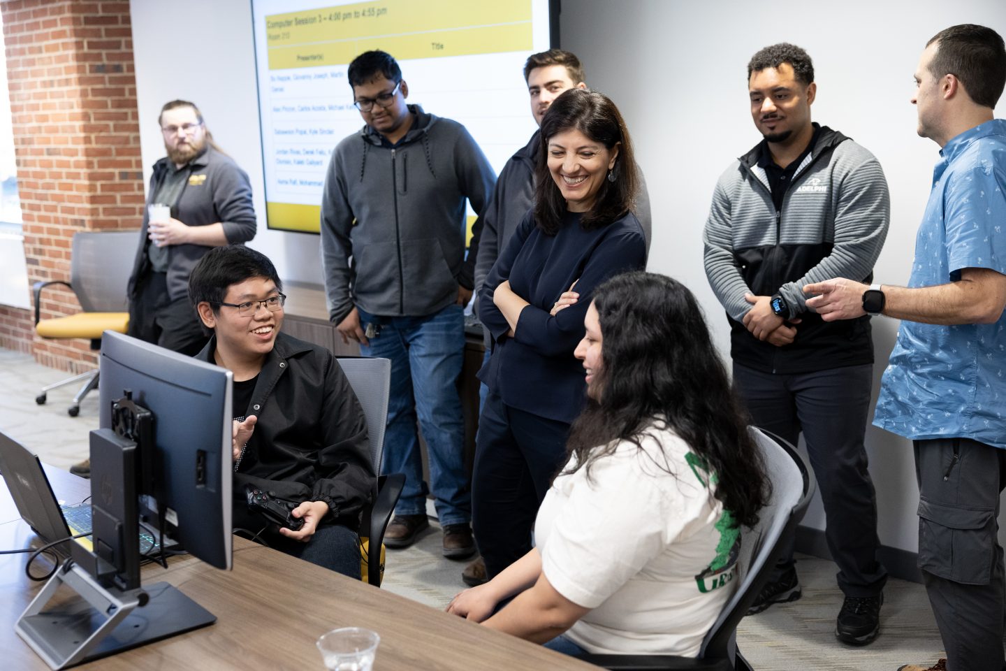 A diverse group of students and adults—two women and six men—are gathered near a computer terminal. A man, seated, and two women, one seated, are in a discussion and smiling, as the others stand by a wall with a large video screen. 